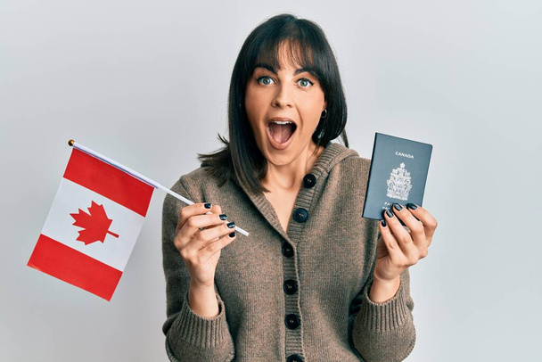 Mujer hispana joven sosteniendo bandera de Canadá y pasaporte celebrando loco y sorprendido por el éxito con los ojos abiertos gritando emocionado.  - Foto, Imagen