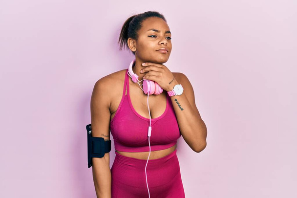 jong Afrikaans amerikaans meisje dragen sportschool kleding en met behulp van koptelefoon aanraken pijnlijke nek, zere keel voor griep, clod en infectie  - Foto, afbeelding