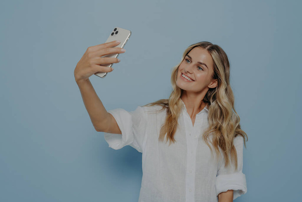 Bella giovane femmina con lunghi capelli biondi tinti che tengono il telefono cellulare e posano per selfie - Foto, immagini