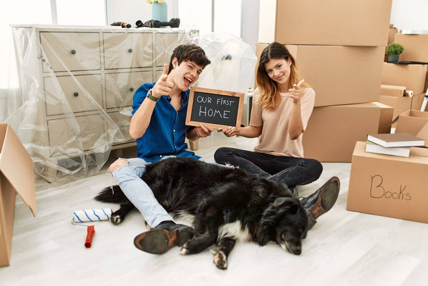 Jeune couple caucasien avec chien tenant notre premier tableau noir à la maison nouvelle pointant les doigts vers la caméra avec un visage heureux et drôle. bonne énergie et vibes.  - Photo, image