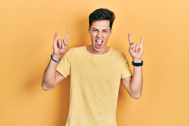 Young hispanic man wearing casual yellow t shirt shouting with crazy expression doing rock symbol with hands up. music star. heavy music concept.  - 写真・画像