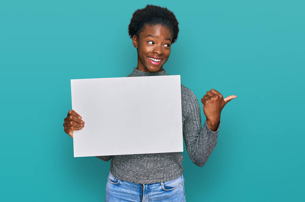 Joven afroamericana americana sosteniendo en blanco banner vacío señalando el pulgar hacia el lado sonriendo feliz con la boca abierta  - Foto, imagen