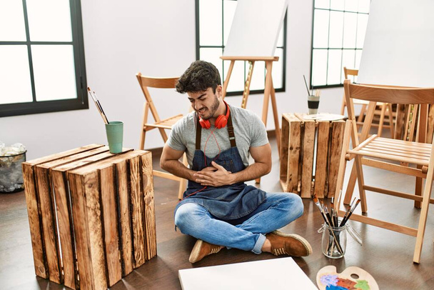Young hispanic man sitting at art studio with hand on stomach because nausea, painful disease feeling unwell. ache concept.  - Valokuva, kuva