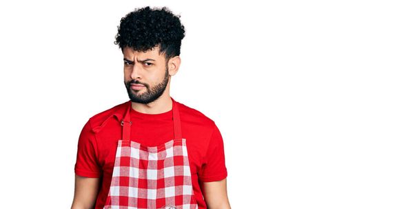 Молодий арабський чоловік з бородою носить кулінарний фартух скептично і нервово, розчаровуючись через проблеми. негативна людина
.  - Фото, зображення