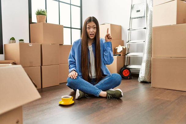 Menina chinesa jovem sentado no chão em nova casa apontando dedo para cima com ideia de sucesso. saiu e feliz. Número um..  - Foto, Imagem