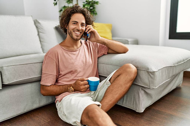 Giovane uomo ispanico bere caffè e parlare sullo smartphone seduto sul pavimento a casa. - Foto, immagini