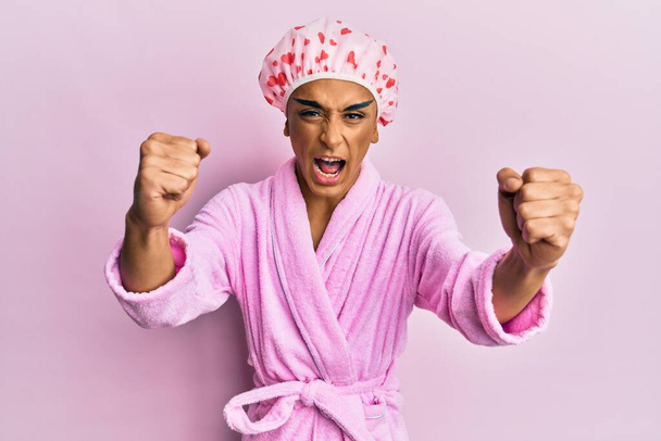 Hispanic man wearing make up wearing shower towel cap and bathrobe angry and mad raising fists frustrated and furious while shouting with anger. rage and aggressive concept.  - 写真・画像