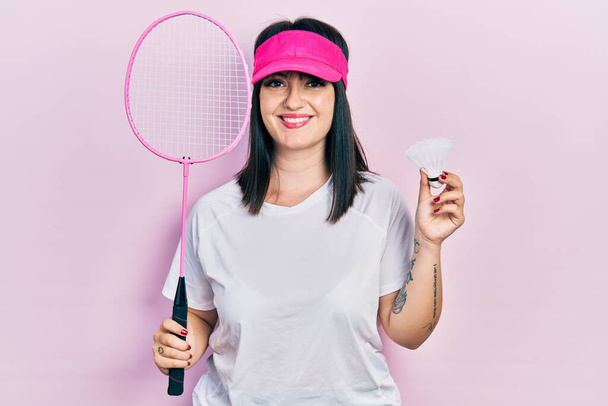 Młoda latynoska kobieta trzymająca rakietę badmintona i wahadłowca uśmiechająca się ze szczęśliwym i chłodnym uśmiechem na twarzy. wykazujące zęby.  - Zdjęcie, obraz