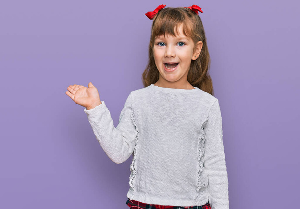 Kis kaukázusi lány gyerek alkalmi ruhát visel mosolygós vidám bemutató és mutató tenyérrel néz a kamerába.  - Fotó, kép