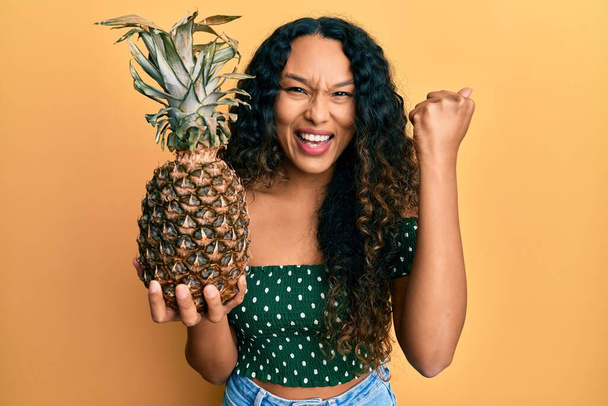Fiatal latin nő tartja ananász sikoltozó büszke, ünnepli győzelem és a siker nagyon izgatott felemelt karral  - Fotó, kép