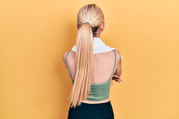 Młoda blondynka ubrana w odzież sportową i ręcznik stojąca tyłem do siebie z skrzyżowanymi ramionami  - Zdjęcie, obraz
