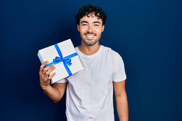 Joven hombre hispano sosteniendo regalo luciendo positivo y feliz de pie y sonriendo con una sonrisa confiada mostrando dientes  - Foto, imagen