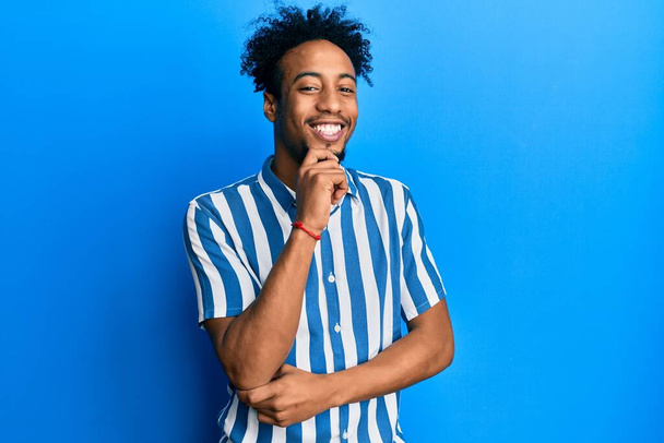 Jeune homme afro-américain barbu portant une chemise rayée décontractée regardant avec confiance la caméra avec le sourire avec les bras croisés et la main levée sur le menton. penser positivement.  - Photo, image