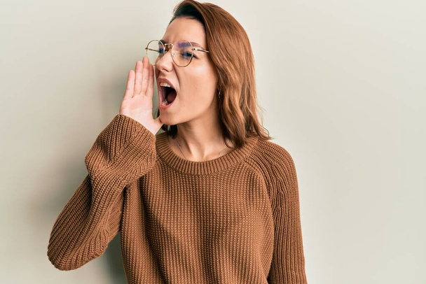 Mujer caucásica joven con ropa casual y gafas gritando y gritando fuerte a un lado con la mano en la boca. concepto de comunicación.  - Foto, imagen