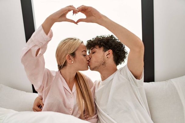 Joven hermosa pareja haciendo símbolo del corazón con las manos sentadas en la cama en casa. - Foto, Imagen