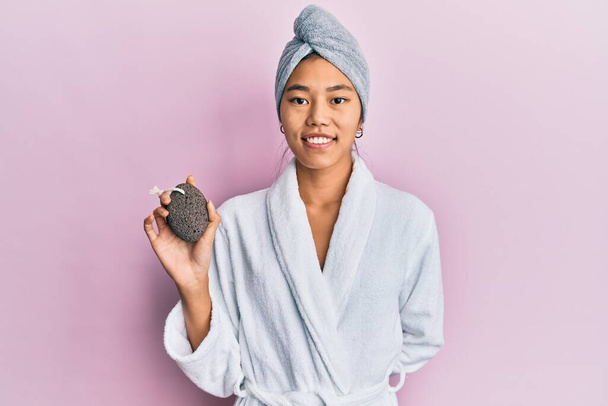 Jeune femme chinoise portant un bonnet de serviette de douche et peignoir tenant pierre ponce regardant positif et heureux debout et souriant avec un sourire confiant montrant des dents  - Photo, image