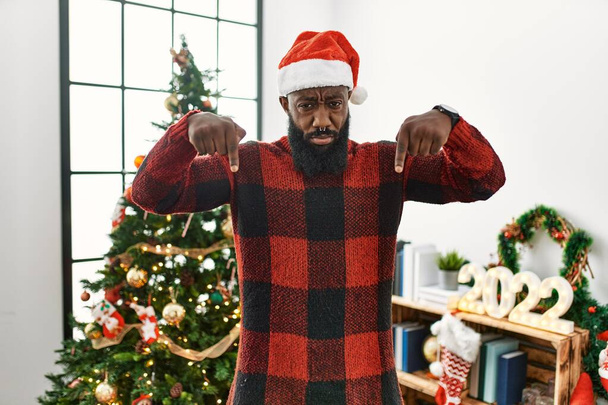 African american man wearing santa claus hat standing by christmas tree pointing down looking sad and upset, indicating direction with fingers, unhappy and depressed.  - Φωτογραφία, εικόνα