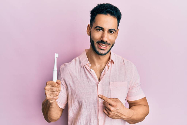 Jovem hispânico segurando escova de dentes elétrica sorrindo feliz apontando com mão e dedo  - Foto, Imagem