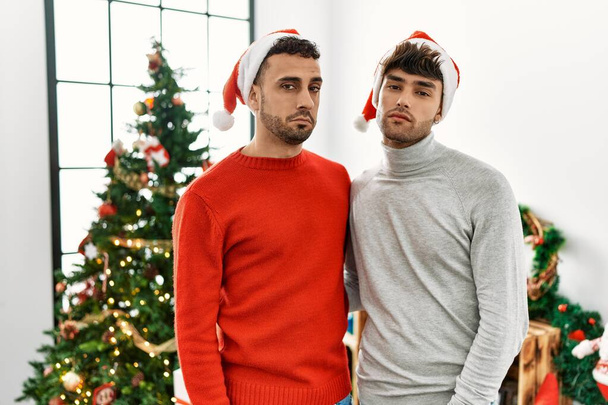 Νεαρό γκέι ζευγάρι στέκεται δίπλα στο χριστουγεννιάτικο δέντρο φορώντας καπέλο που δείχνει νυσταγμένο και κουρασμένο, εξαντλημένο από την κούραση και το μεθύσι, τεμπέλικα μάτια το πρωί.  - Φωτογραφία, εικόνα