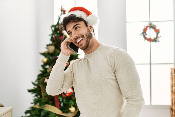 Mladý hispánec nosí vánoční klobouk mluví na smartphone doma. - Fotografie, Obrázek