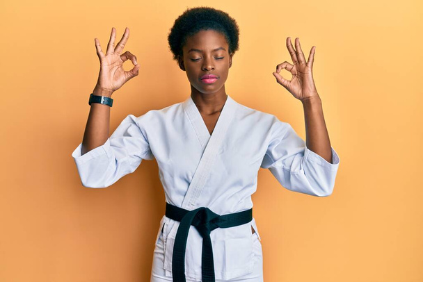 Mladá Afroameričanka v karate kimonu a černém opasku odpočívá a usmívá se se zavřenýma očima a prsty dělá meditační gesto. koncepce jógy.  - Fotografie, Obrázek