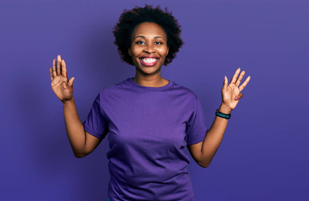 Afroamerykanka z afro włosy na sobie casual fioletowy t koszulka pokazując i wskazując palcem numer dziewięć uśmiechając się pewny siebie i szczęśliwy.  - Zdjęcie, obraz