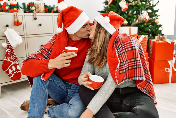 Молода пара носить різдвяний капелюх, покритий ковдрою. Сидячи на підлозі п'є каву вдома
. - Фото, зображення