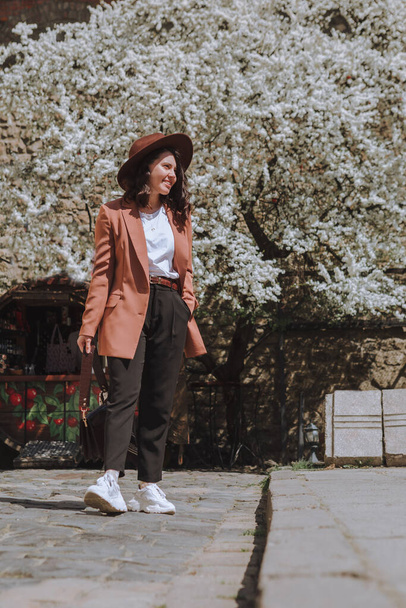 улыбающаяся красивая женщина в куртке и шляпе прогуливаясь по весенней улице белого цветущего дерева на заднем плане - Фото, изображение