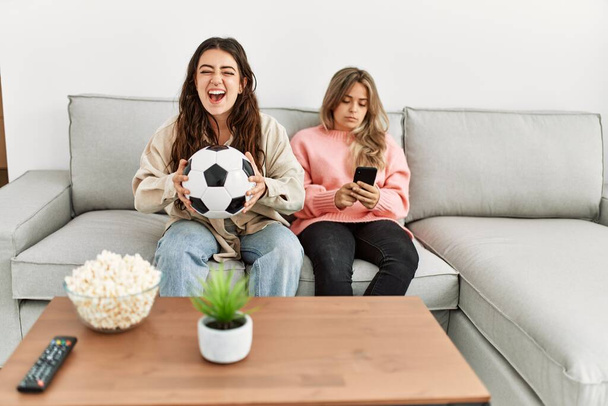 Girl smiling happy watching soccer match and her girlfriend boring using smartphone at home. - Fotografie, Obrázek