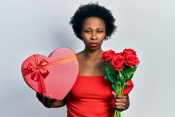 Mujer afroamericana joven sosteniendo regalo de aniversario y ramo de flores escéptico y nervioso, frunciendo el ceño molesto debido a un problema. persona negativa.  - Foto, imagen
