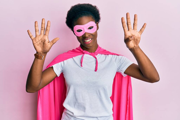 Young african american girl wearing superhero mask and cape costume showing and pointing up with fingers number nine while smiling confident and happy.  - Photo, Image