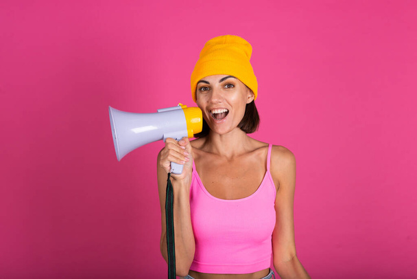 Brillante retrato de mujer en sombrero amarillo sobre fondo rosa gritando en megáfono - Foto, Imagen
