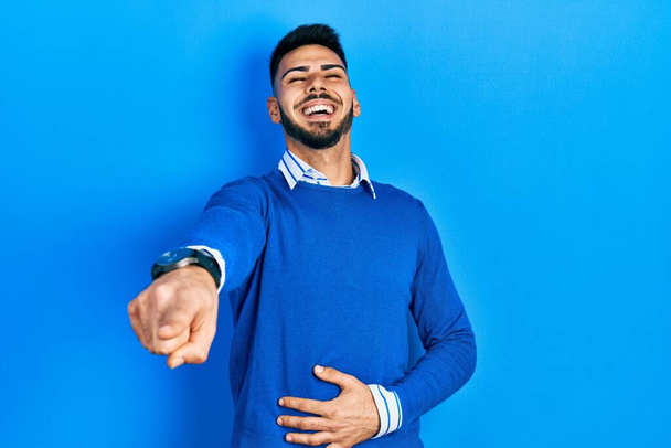 Mladý hispánec s plnovousem v neformálním modrém svetru, směje se ti, ukazuje prstem na kameru s rukou na těle, výraz studu  - Fotografie, Obrázek