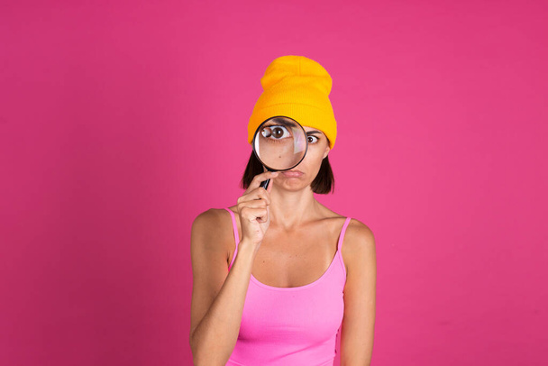 Bright portrait of woman in yellow hat on pink background with magnifier angry confused - Фото, изображение