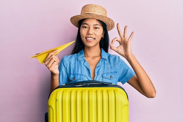 Νεαρή Κινέζα γυναίκα κρατώντας τσάντα καμπίνα και χάρτινο αεροπλάνο κάνει ok υπογράψει με τα δάχτυλα, χαμογελώντας φιλικό gesturing εξαιρετικό σύμβολο  - Φωτογραφία, εικόνα