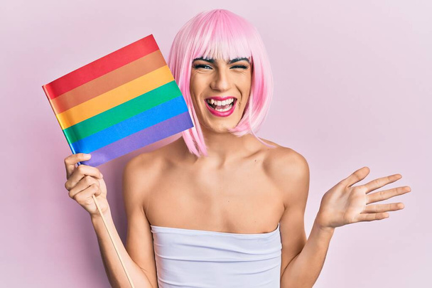 Young man wearing woman make up holding rainbow lgbtq flag wearing pink wig celebrating achievement with happy smile and winner expression with raised hand  - Φωτογραφία, εικόνα