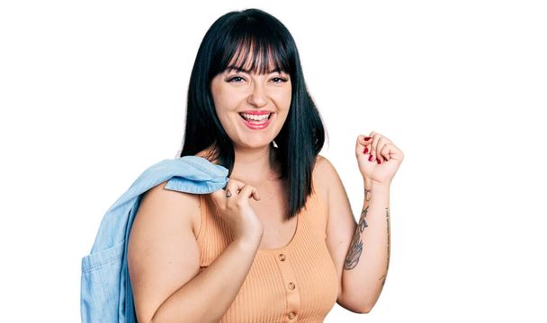 Young hispanic plus size woman wearing holding denim jacket screaming proud, celebrating victory and success very excited with raised arm  - 写真・画像