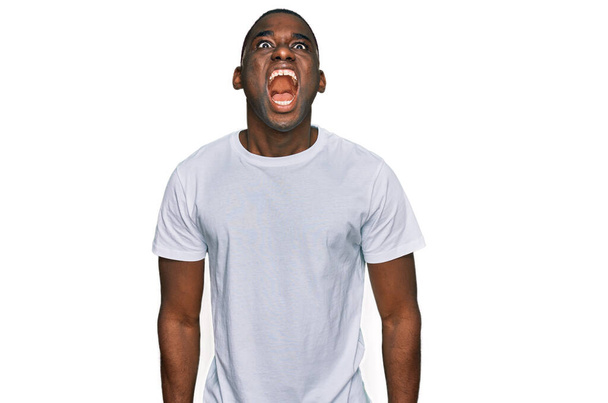 Jeune homme afro-américain portant un t-shirt blanc décontracté en colère et fou hurlant frustré et furieux, criant de colère. rage et concept agressif.  - Photo, image