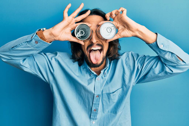 Young hispanic man holding canned food over eyes sticking tongue out happy with funny expression.  - Photo, Image