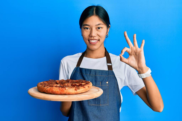 Garson önlüğü giyen genç Çinli kadın İtalyan pizzası tutuyor. Parmaklarıyla imza atıyor, gülümsüyor. Mükemmel bir sembol.  - Fotoğraf, Görsel