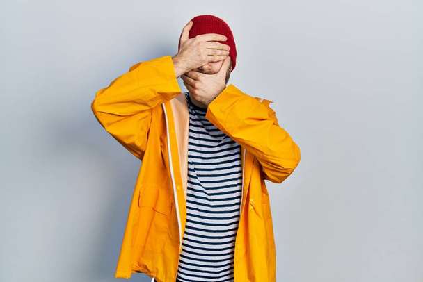 Caucasian man with beard wearing yellow raincoat covering eyes and mouth with hands, surprised and shocked. hiding emotion  - Foto, Bild