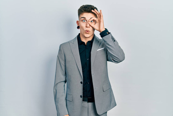 Young caucasian boy with ears dilation wearing business jacket doing ok gesture shocked with surprised face, eye looking through fingers. unbelieving expression.  - Фото, изображение