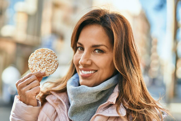 Junge hispanische Frau lächelt glücklich und hält Reiskuchen in der Stadt. - Foto, Bild