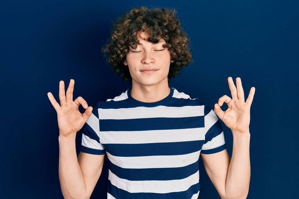 Jóképű fiatalember alkalmi csíkos pólót visel lazítson és mosolyogjon csukott szemmel meditációs gesztussal az ujjaival. jóga koncepció.  - Fotó, kép