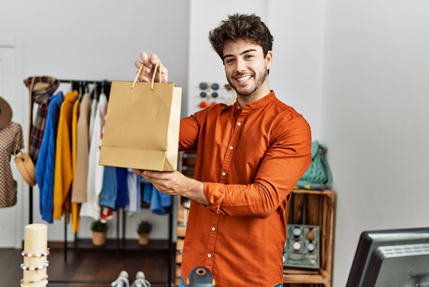 Joven comerciante hispano sonriendo feliz sosteniendo bolsa de compras en tienda de ropa. - Foto, Imagen