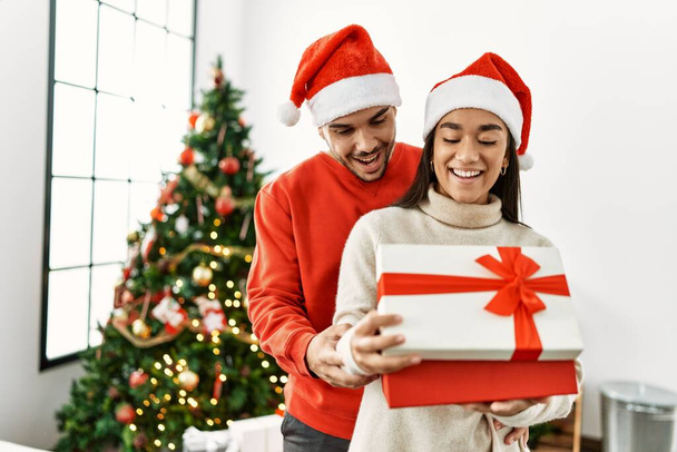 Joven pareja latina sonriendo feliz y abrazando unboxing regalo de Navidad en casa. - Foto, Imagen