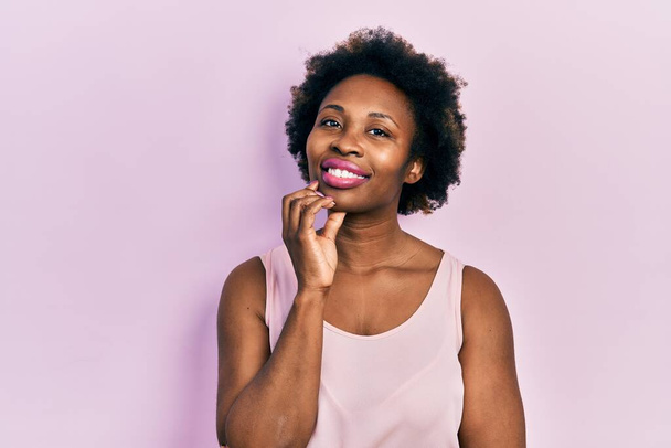 Young african american woman wearing casual sleeveless t shirt looking confident at the camera with smile with crossed arms and hand raised on chin. thinking positive.  - Photo, Image