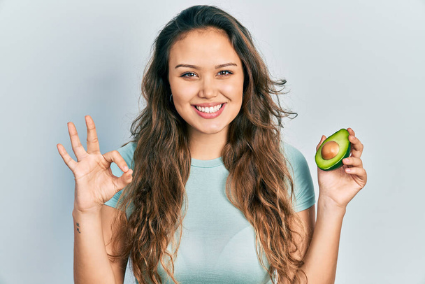 Young hispanic girl holding middle avocado doing ok sign with fingers, smiling friendly gesturing excellent symbol  - Φωτογραφία, εικόνα