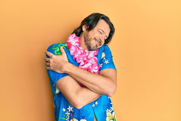 Moyen âge bel homme portant chemise d'été et lei hawaïen se serrant dans ses bras heureux et positifs, souriant confiant. amour de soi et soins de soi  - Photo, image