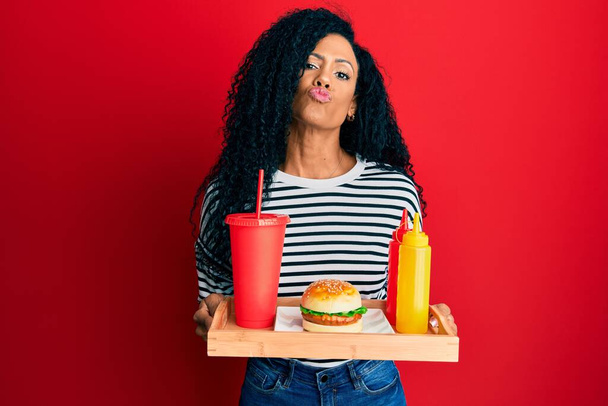 Afro-Amerikaanse vrouw van middelbare leeftijd die een lekkere klassieke hamburger eet, kijkend naar de camera die een kus blaast, heerlijk en sexy. liefde uitdrukking.  - Foto, afbeelding